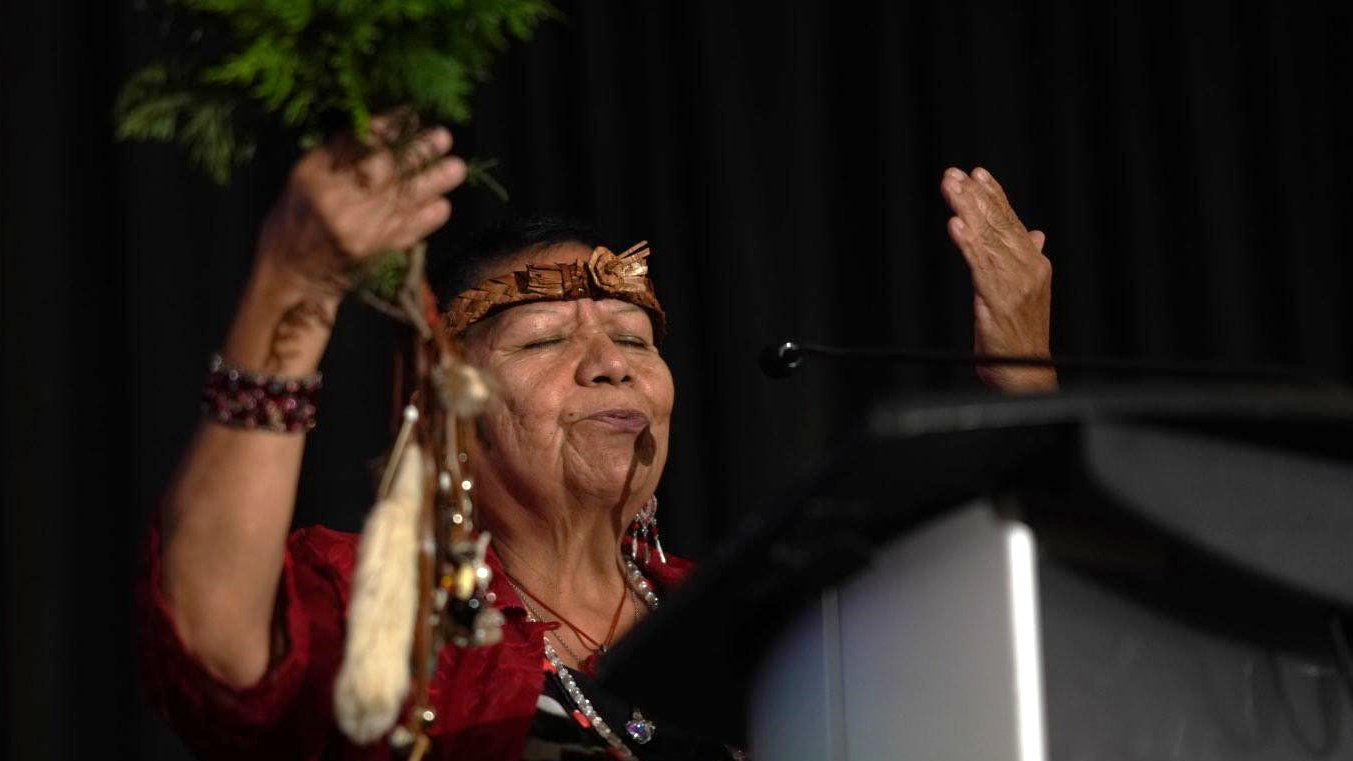 Elder Roberta Price leading an opening prayer at Putting Patients First 2023, with hands raised and dressed in regalia in front of a podium.