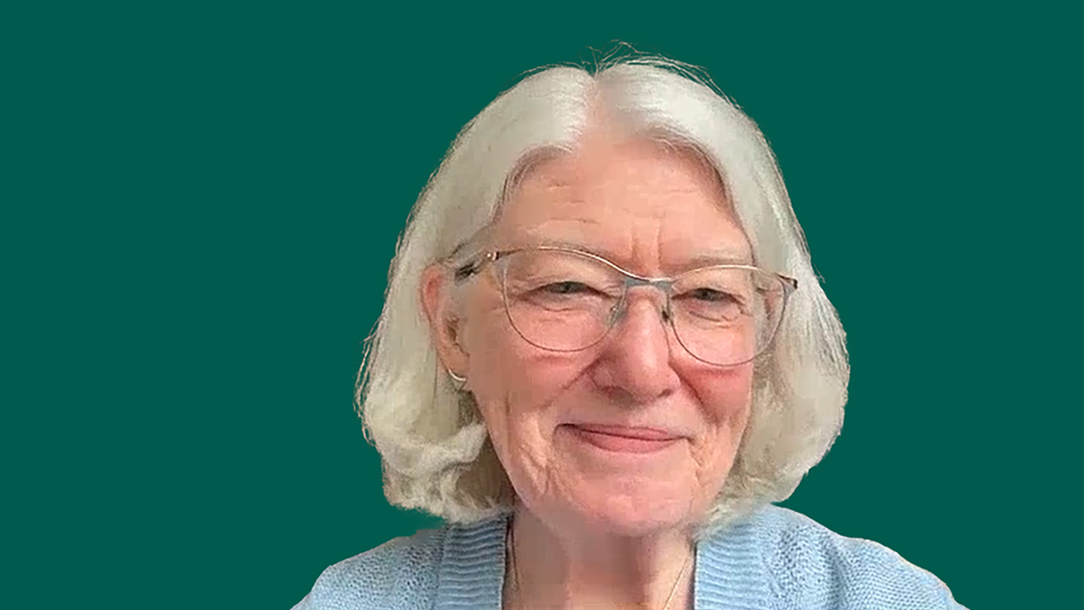 A women with white hair, glasses and light blue shirt. The background is green.