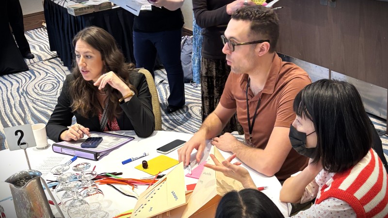 Conference attendees sit at a table with folded origami hats on it. The hats have words that describe different roles that the attendees play in their lives, like "son."