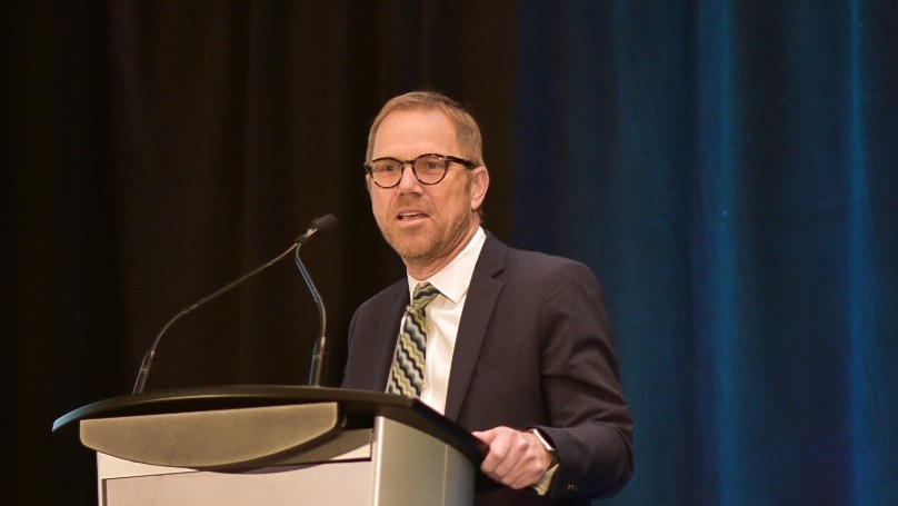 Dr. Robert Reid dressed formally, speaking energetically at a podium on a stage, against dark curtains.