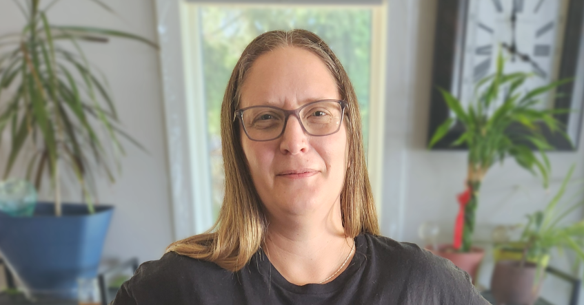 Patient partner Christina LaBerge, smiling. She seems to be standing in her home. A clock, a window and many plants can be seen behind her.