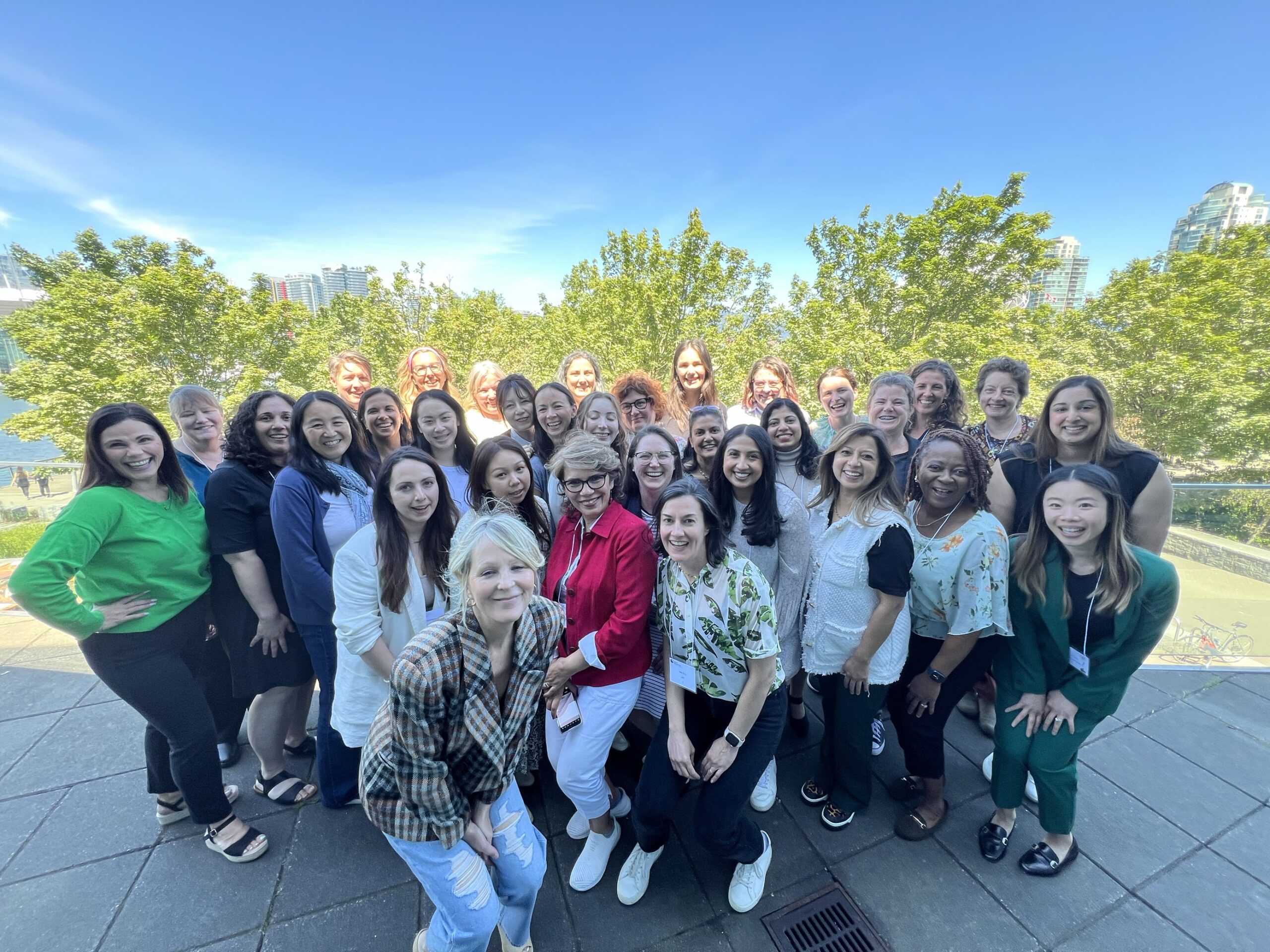 Group photo of the 2024 REBA retreat attendees gathered outdoors with a bright blue sky and trees in the background. The team is smiling and posing together, wearing a variety of colorful outfits.