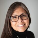 Headshot of Asma-na-hi Antoine smiling broadly, with a soft grey background behind her.