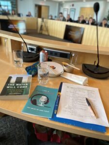Books about podcasting and podcasting notes ontop of a wooden podium. The books are "The Water Will Come" and "A Guide to Academic Podcasting." A crowd is visible behind the podium.